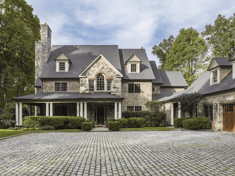 Elegant stone home with gray Enviroshake roof and cobblestone driveway surrounded by trees.