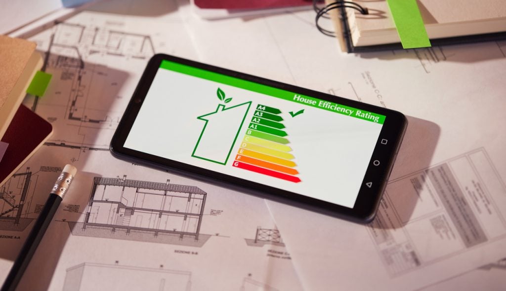 Energy efficiency screen on cellphone leaning on drawings and blueprints of a new house. Architect's desk with plans on it and a cell phone with an energy efficiency graph. Renewable energy, renovation, energy class and eco house concept.